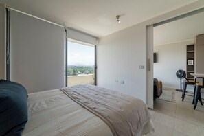 Ein Schlafzimmer mit einem Bett, einem Schreibtisch und einem Sessel, mit Blick auf die Stadt durch ein großes Fenster.