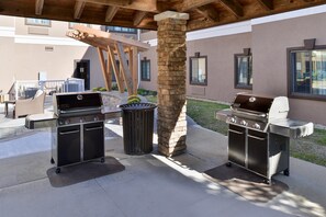 Een buitenpatio met twee barbecues, een vuilnisbak en een houten pergola.