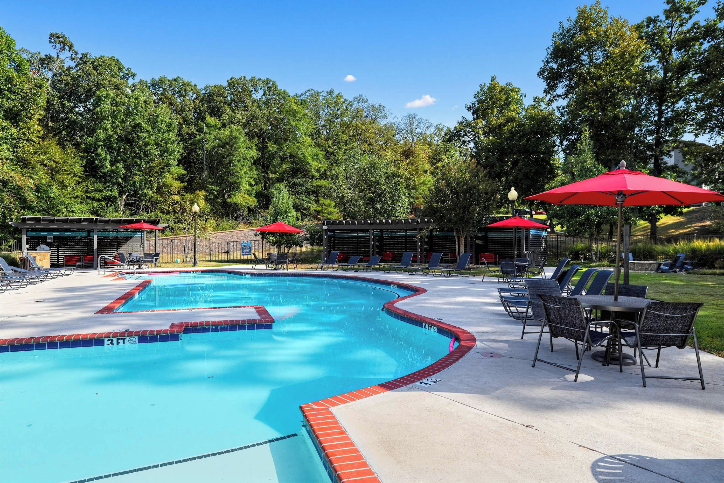 Ein Swimmingpool mit roter Umrandung, umgeben von Liegestühlen und Sonnenschirmen, vor einer Baumkulisse.