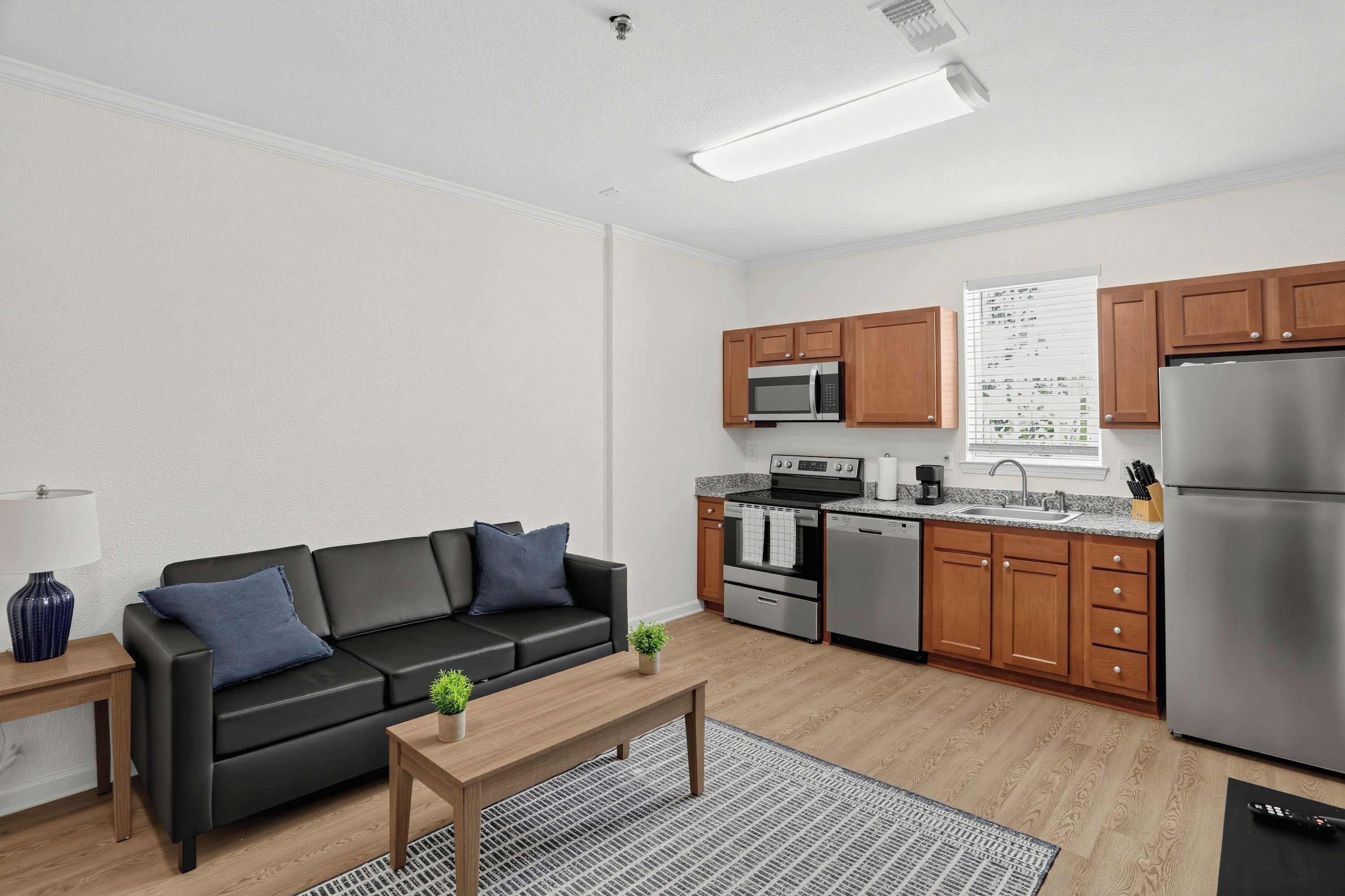 A modern living room with a black leather sofa, a wooden coffee table, and a kitchen area featuring stainless steel appliances and wooden cabinets.