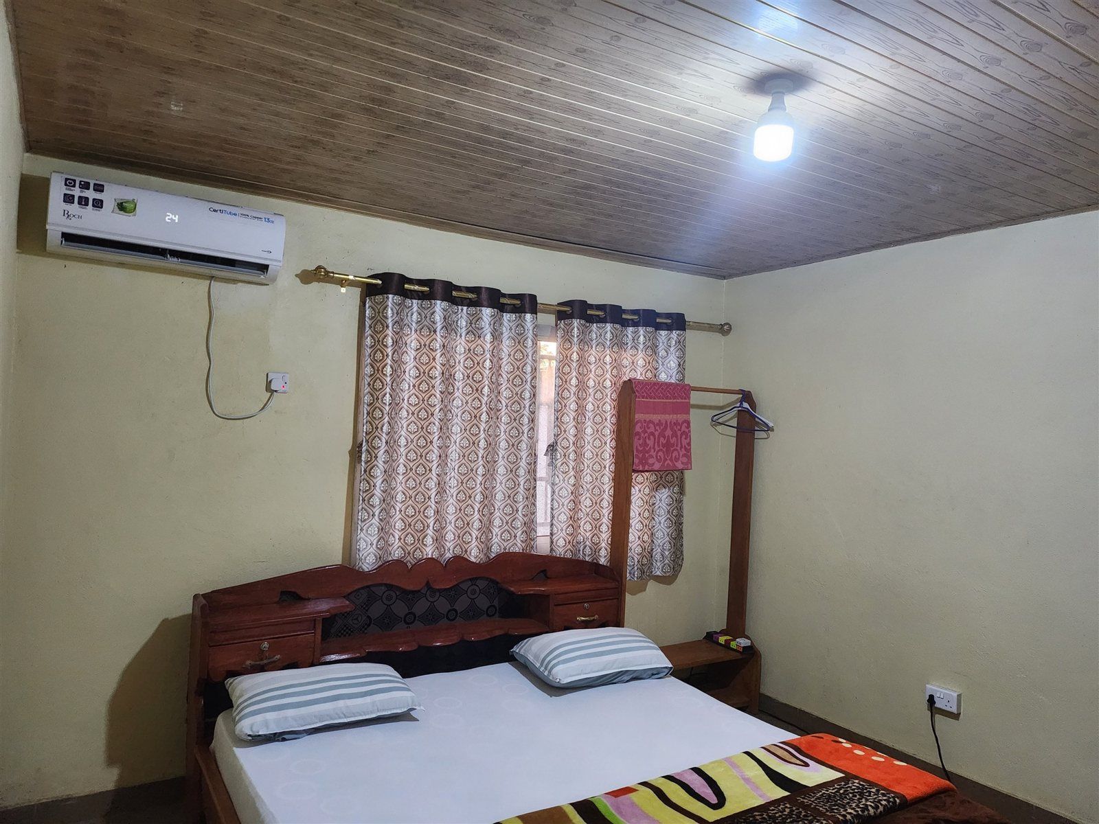 Un dormitorio con techo de madera, una cama con dos almohadas, una mesita de noche, una lámpara colgante y un aire acondicionado de pared.