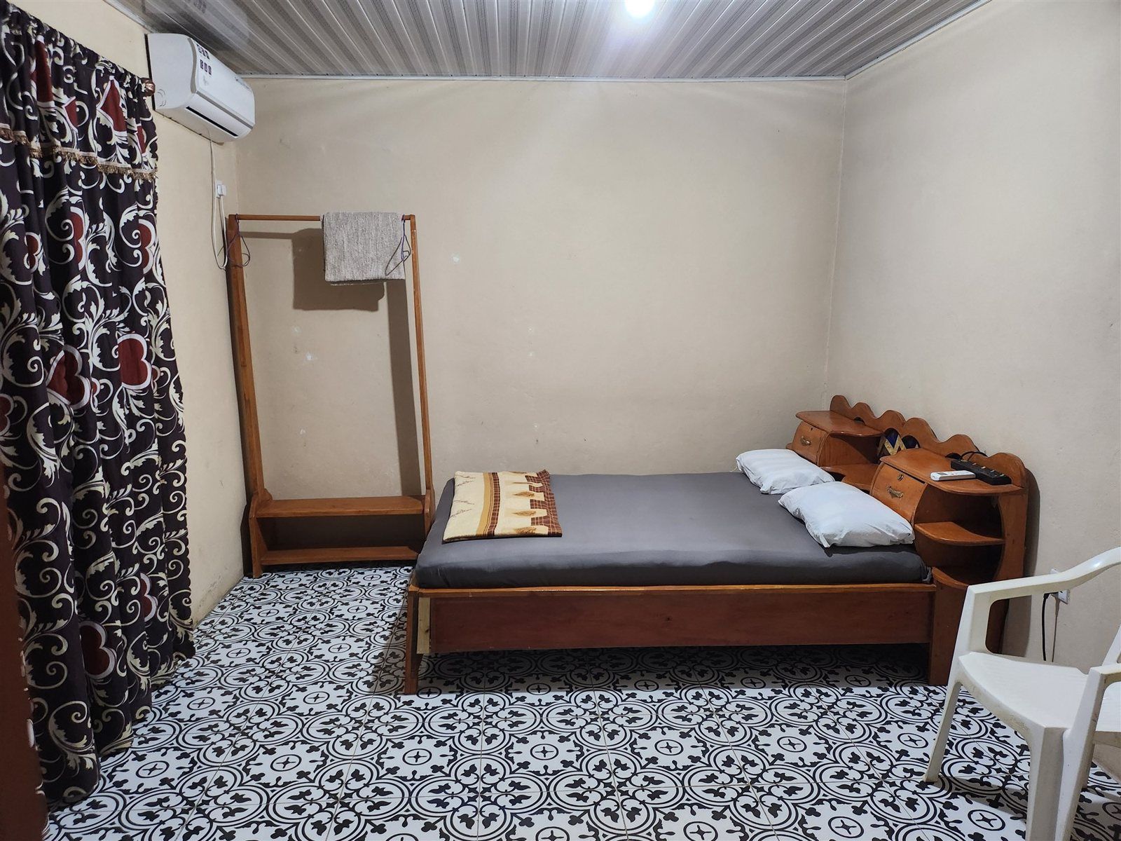 A small, simple room with a bed, a wooden nightstand, a chair, a patterned floor, and a curtain.