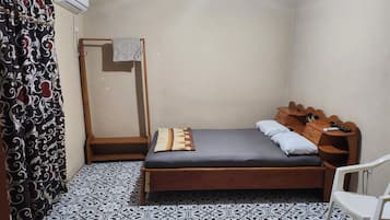 A small, simple room with a bed, a wooden nightstand, a chair, a patterned floor, and a curtain.