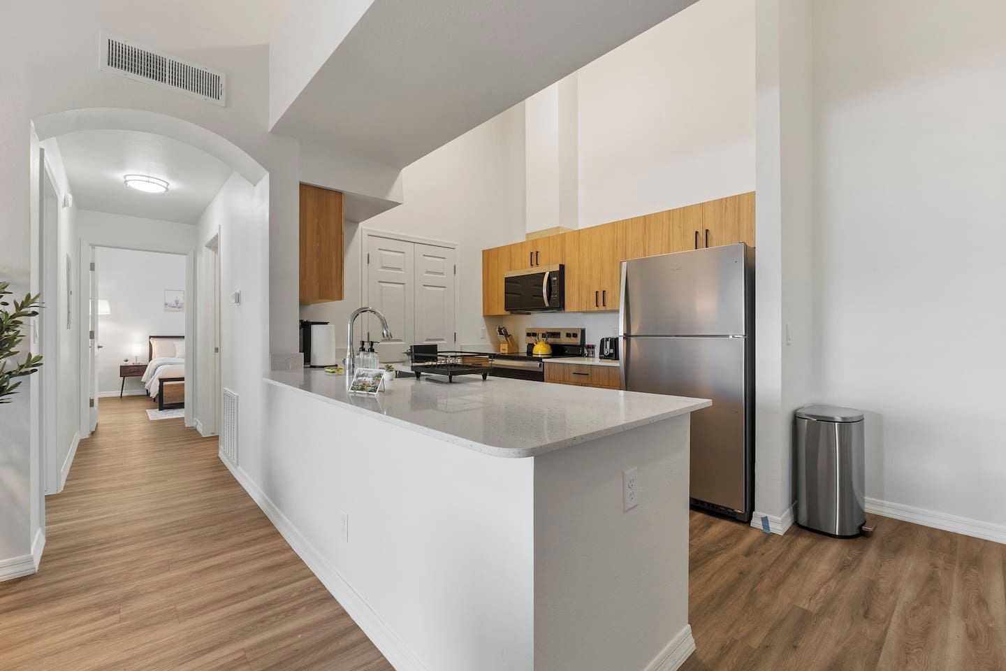 A modern kitchen with a central island, wooden cabinets, stainless steel appliances, and a hallway leading to a bedroom.