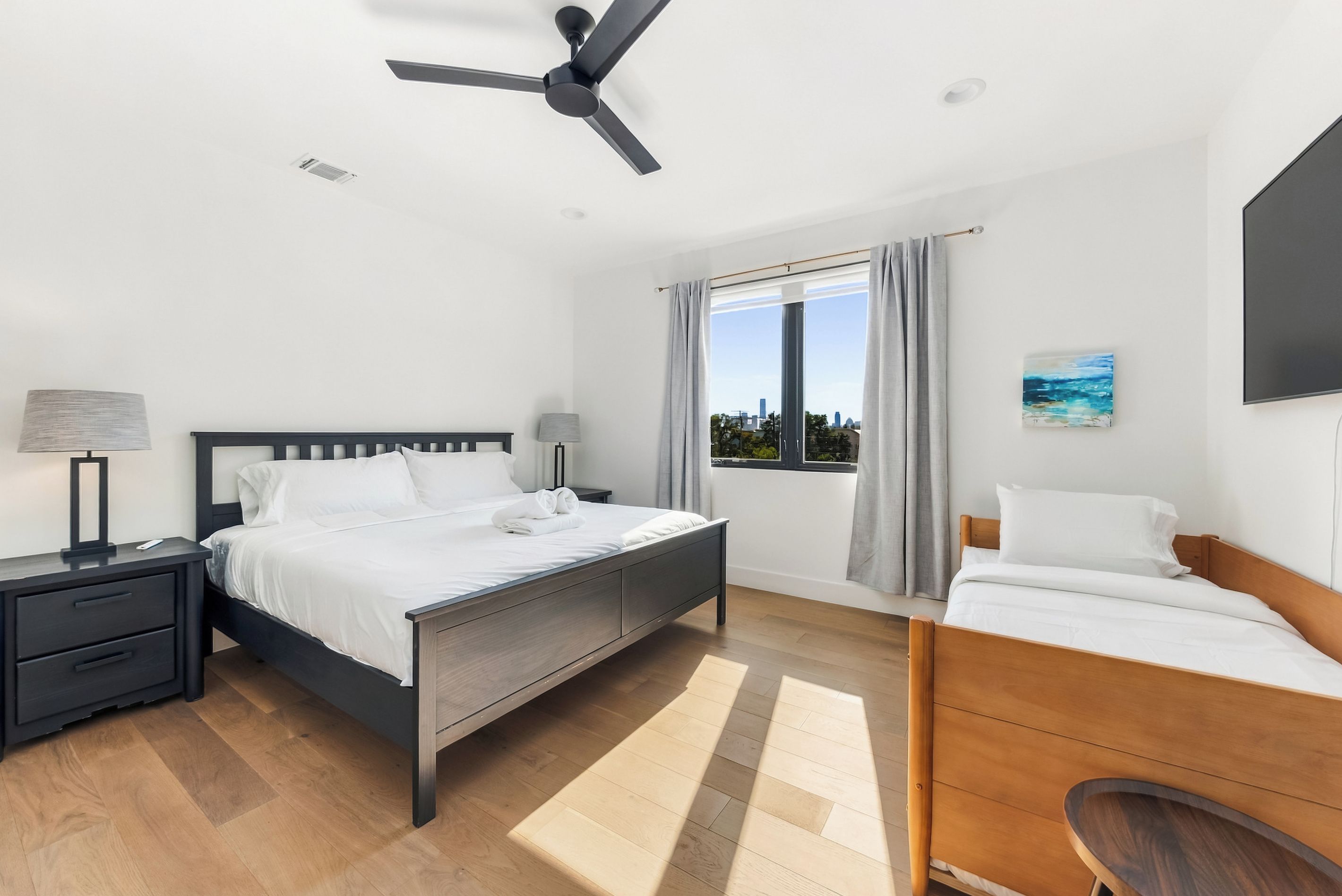 A hotel room with a bed, bedside tables, a desk, a chair, a ceiling fan, a window with curtains, and a painting on the wall.