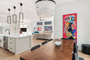 A modern kitchen with white cabinets and a central island, a dining area with a wooden table and chairs, and a colorful abstract painting on the wall. - East Austin Skyline & Pool House ~Backyard BBQ~ (Austin)
