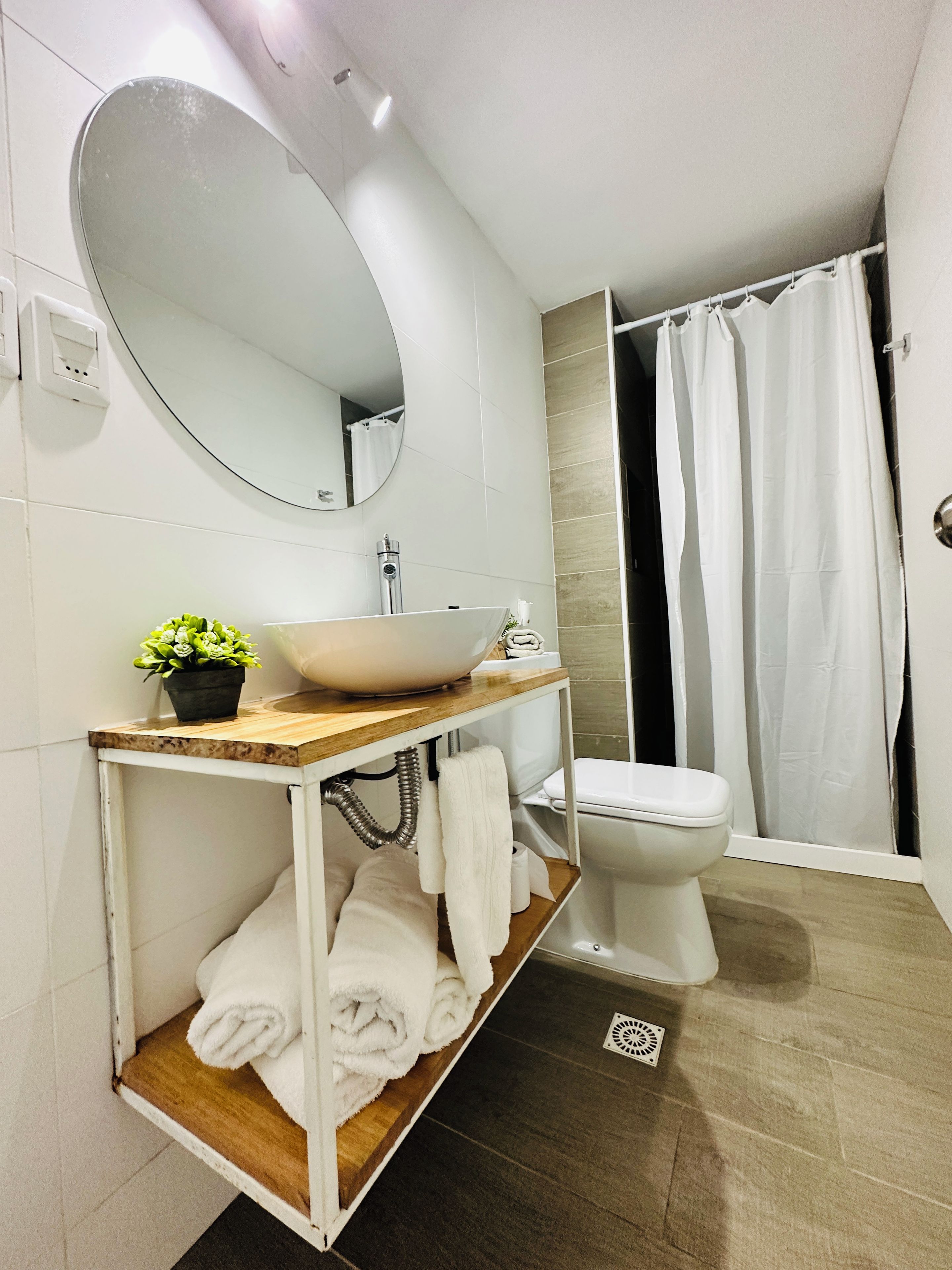 A modern bathroom with a white sink, mirror, and toilet. There is a shower curtain, a potted plant, and a shelf with towels.