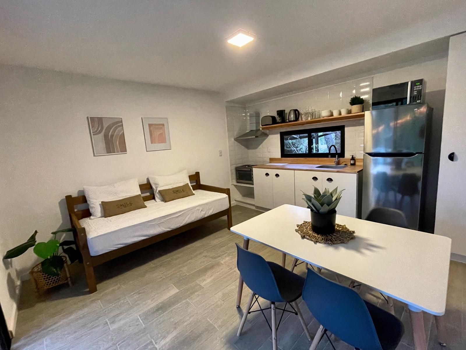 Cocina compacta con refrigerador, microondas y estufa. Hay una mesa de comedor con sillas y una planta pequeña. Un sofá con almohadas está apoyado contra la pared.