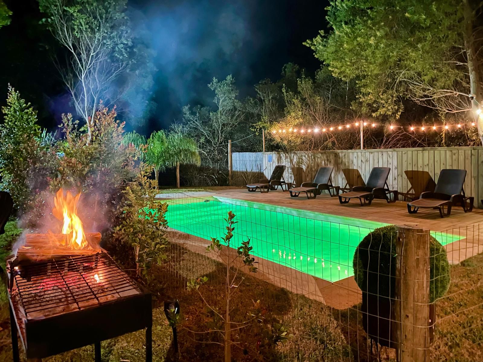 Escena nocturna al aire libre con piscina, sillas de descanso y una parrilla.