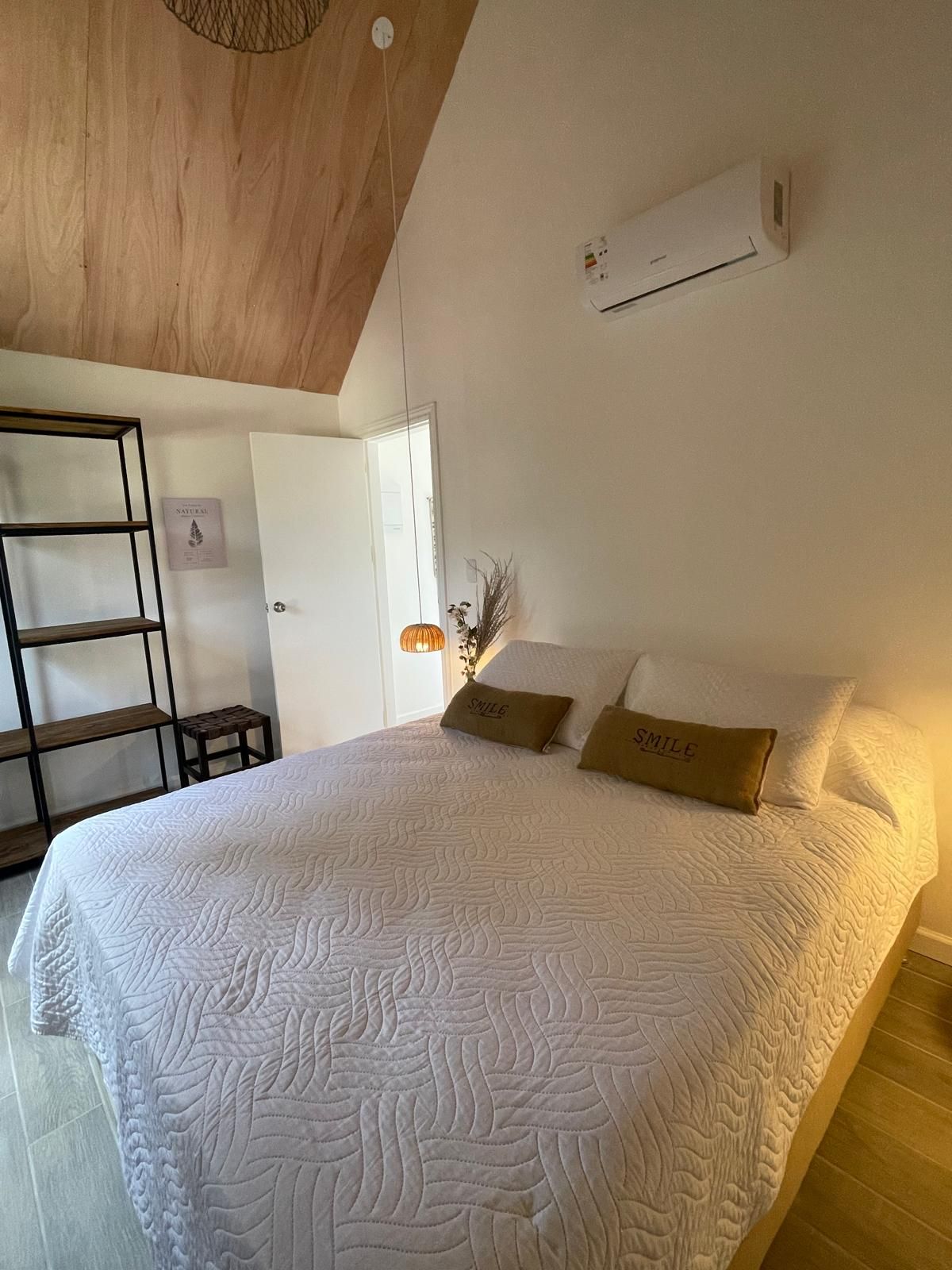 Une chambre avec un grand lit, un plafond en bois, un appareil de climatisation, une petite table et une étagère fixée au mur.
