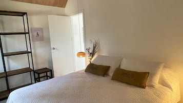 A bedroom with a large bed, a wooden ceiling, an air conditioning unit, a small table, and a wall-mounted shelf.