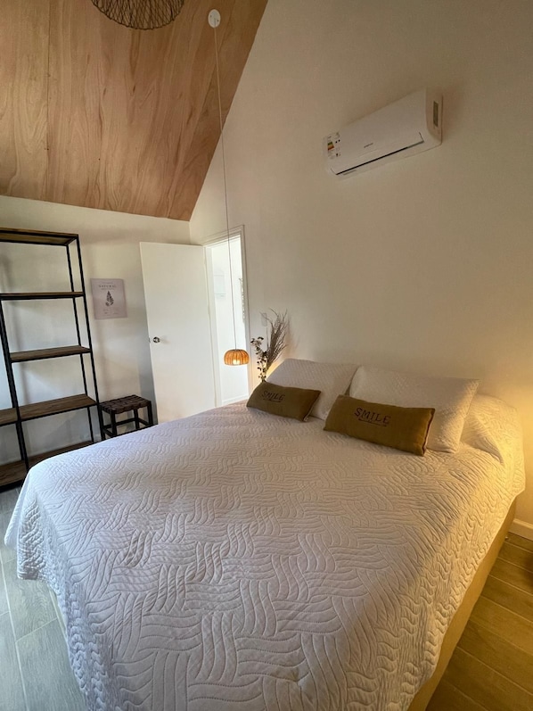 A bedroom with a large bed, a wooden ceiling, an air conditioning unit, a small table, and a wall-mounted shelf. - Villa Mora (Punta Colorada)