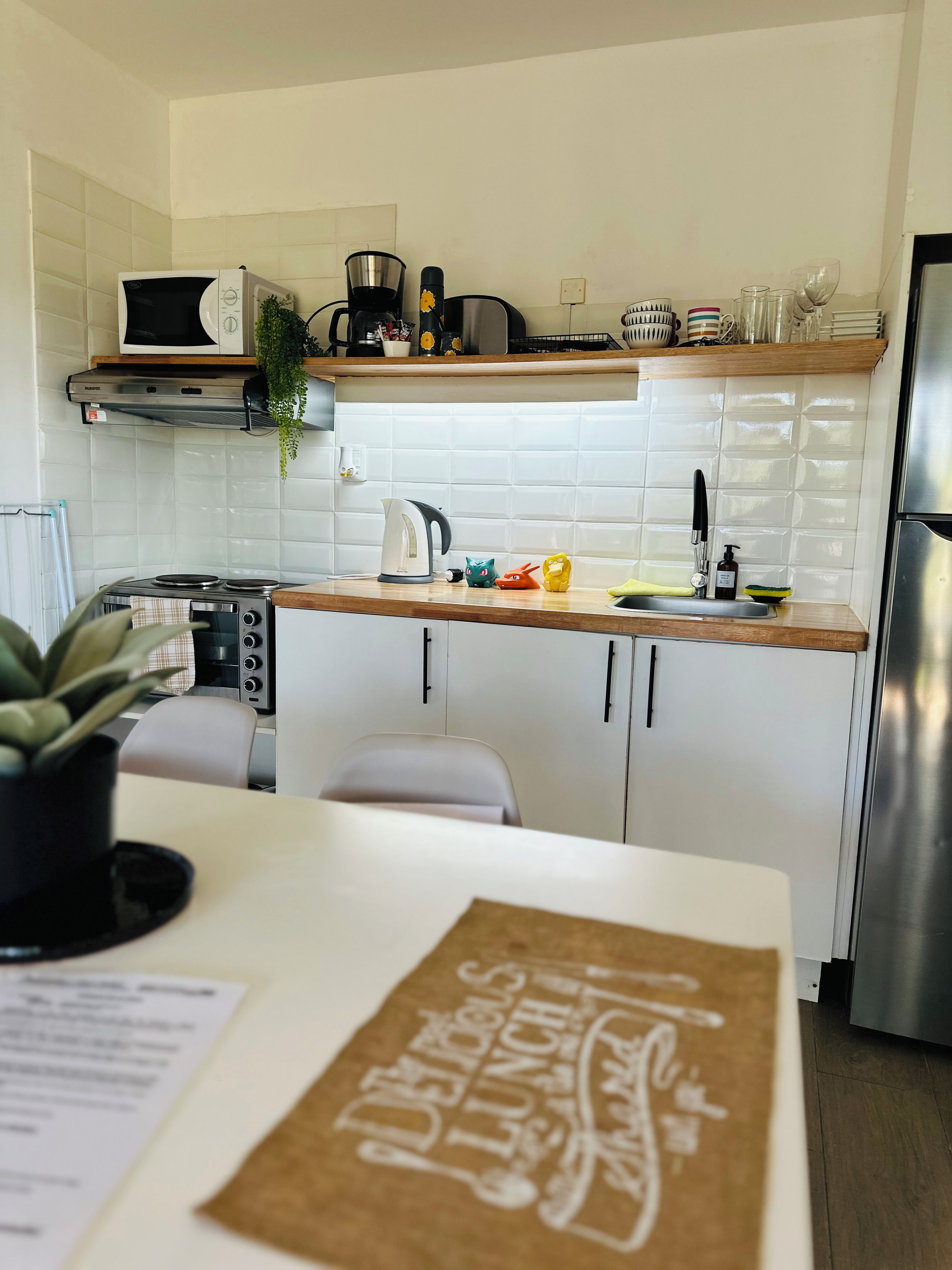 Une cuisine moderne avec des meubles blancs, un four à micro-ondes, une bouilloire et une machine à café. On y trouve un évier avec un robinet, ainsi qu’un plan de travail sur lequel sont disposés divers ustensiles de cuisine.