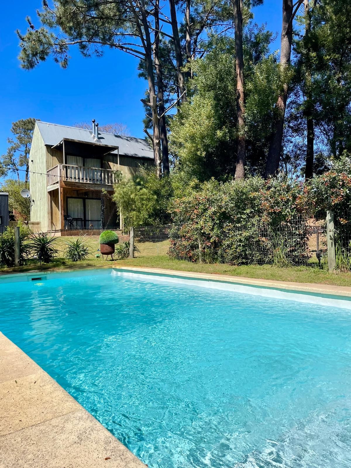 A swimming pool with a wooden deck, surrounded by trees and a house with a balcony.