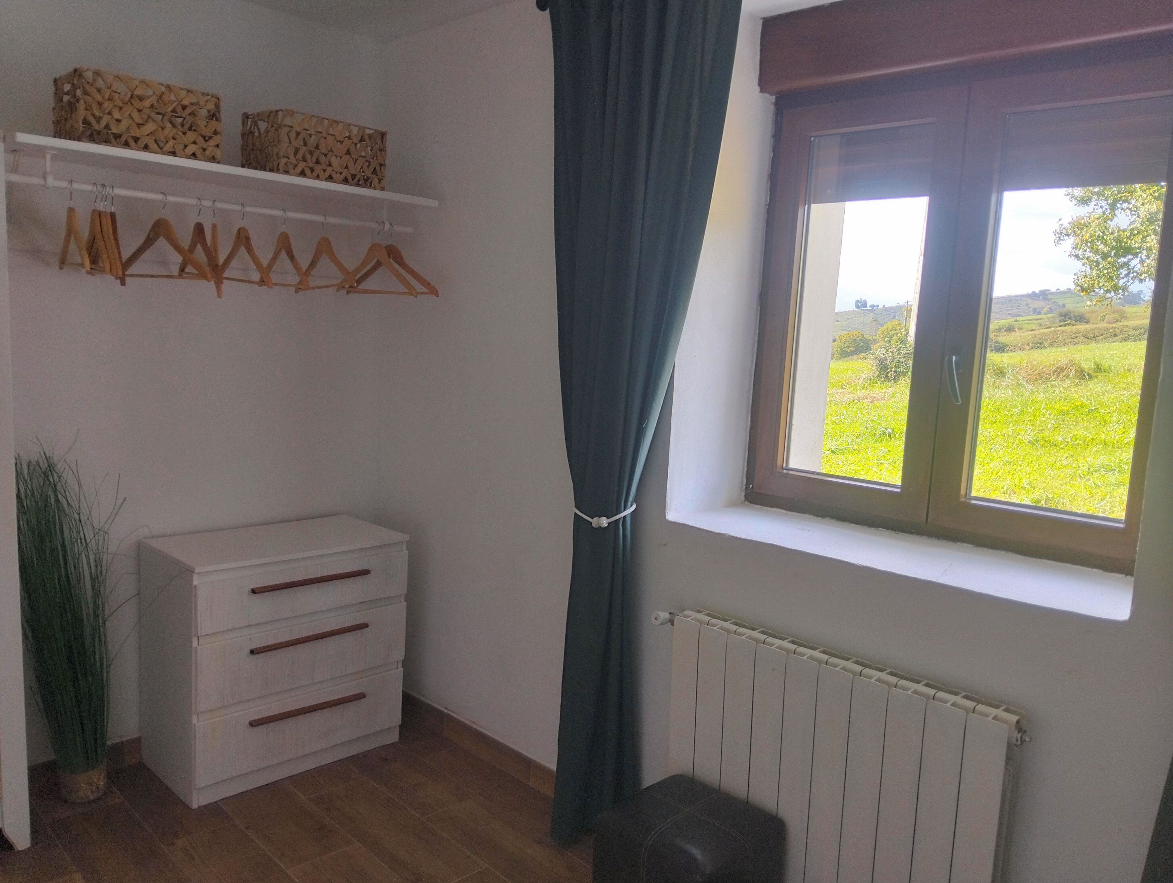 Una stanza con una cassettiera bianca, grucce di legno su un supporto a parete, una finestra che si affaccia su un campo verde e un termosifone.