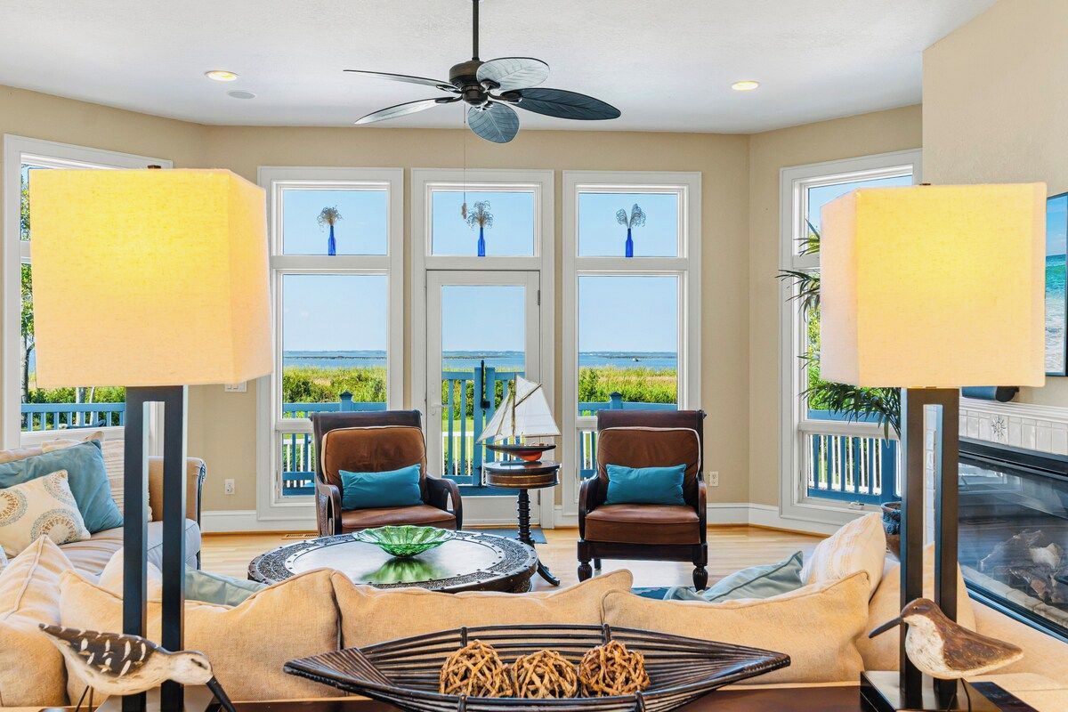 A spacious living room with a view of the ocean, featuring a ceiling fan, two chairs, a coffee table, and a fireplace.