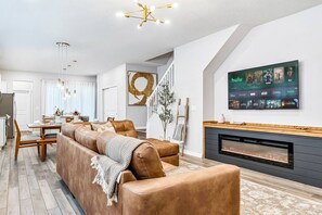 A modern living room with a fireplace, a mounted TV displaying streaming services, and a dining area in the background. - Dino + Gamer Kids Rooms · Private Pool Resort (Clermont)