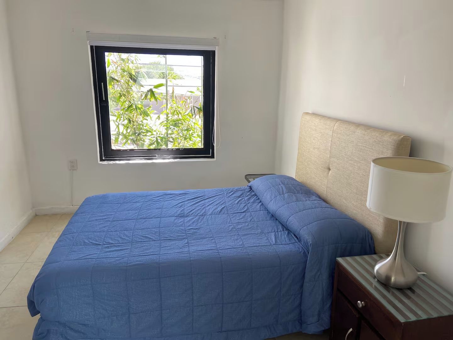 A bedroom with a bed, a nightstand, a lamp, and a window with a view of greenery.