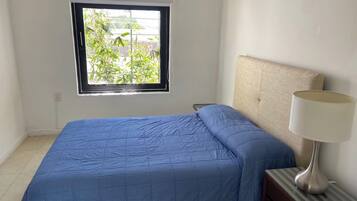 A bedroom with a bed, a nightstand, a lamp, and a window with a view of greenery.