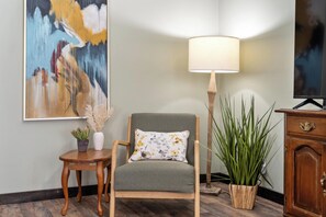 Ein Zimmer mit einem grauen Sessel, einem kleinen Holztischchen mit einem Blumentopf, einer Stehlampe und einem Schrank mit einem Fernseher.