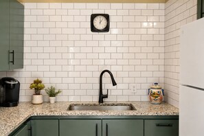 Eine Küche mit weißen, im Ziegelstil gehaltenen Fliesen, einem schwarzen Wasserhahn, einer Keurig-Kaffeemaschine, Blumentöpfen und einem farbenfrohen Keramikbehälter.