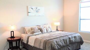 A bedroom with a large bed, two bedside tables, a nightstand, a wall-mounted lamp, and a window with blinds.