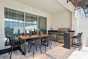 An outdoor patio with a wooden table and chairs, a grill, and a view through large windows. - Luxury 6BR Home with Private Pool at Solara (Kissimmee)