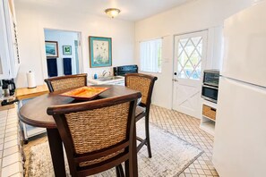 A dining area with a round table and four wicker chairs, a kitchen with a double oven, and a white refrigerator. - Kaneohe Bay 2BR - Oceanfront Pier & Kayaks - Wi-Fi (Kaneohe)