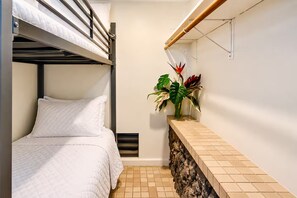 En køjeseng med hvidt quiltet sengetæppe og puder, en lille plante på en hylde, og et flisegulv.