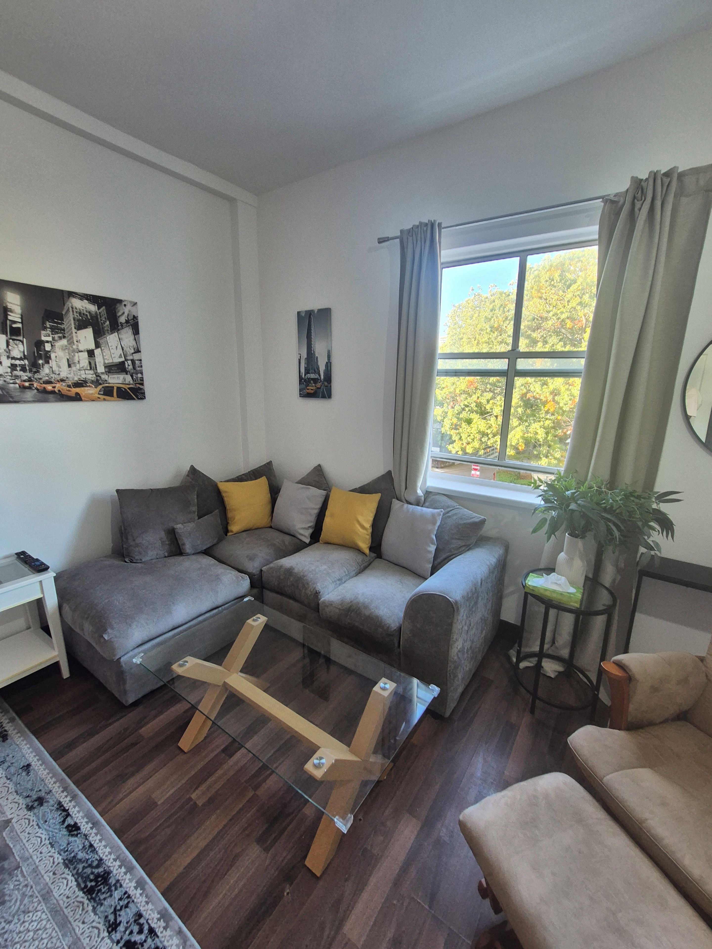 Una sala de estar moderna con un sofá seccional, una mesa de centro de vidrio, una mesita auxiliar y una ventana con cortinas.