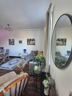 Una sala de estar con sofá, sillón, mesa de centro y comedor con mesa de vidrio y sillas. Hay tres cuadros en la pared, un espejo y una planta en maceta.