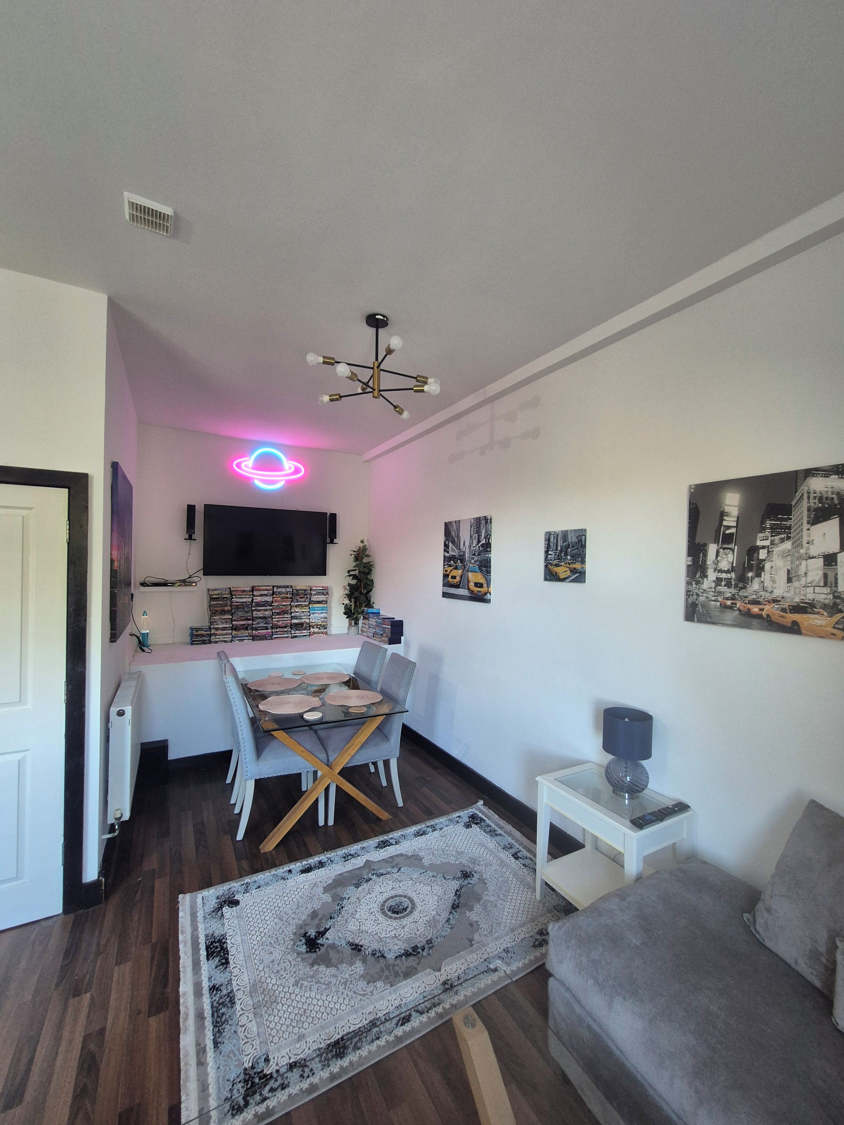 A living room with a dining area, a flat-screen TV mounted on the wall, a ceiling light fixture, and a patterned rug on the floor.