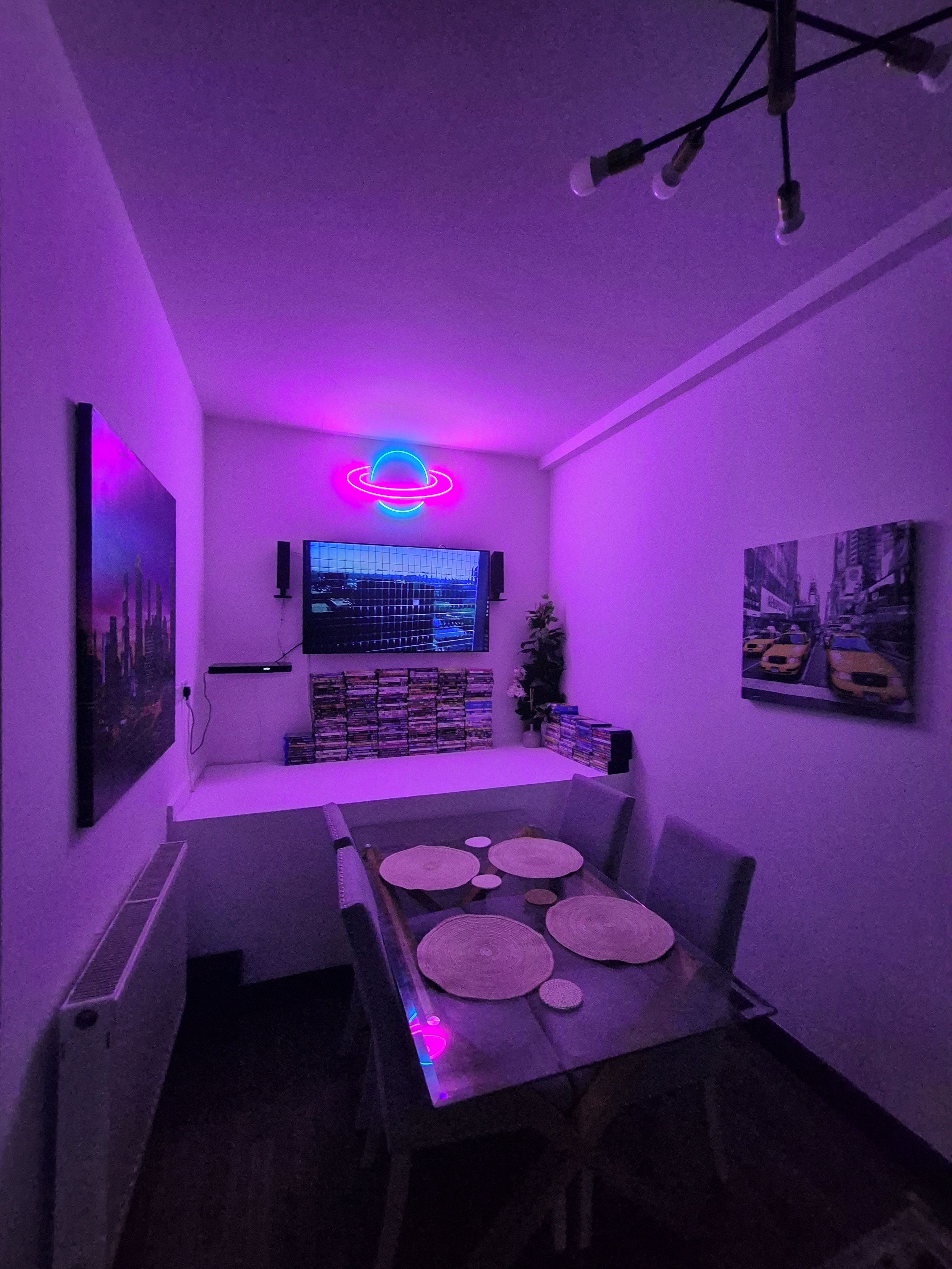 A room with a neon sign, a flat-screen TV, a bookshelf, and a dining area with a table and chairs.
