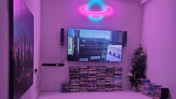 A room with a flat-screen TV mounted on the wall, a stack of DVDs, a small plant, and a neon light.
