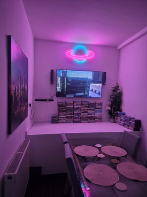 Una stanza con una TV a schermo piatto montata al muro, una pila di DVD, una piccola pianta e una luce al neon.