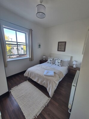 Un dormitorio con un ventanal grande, una cama con ropa de cama blanca, dos mesitas de noche con lámparas, y una cita inspiradora enmarcada en la pared.