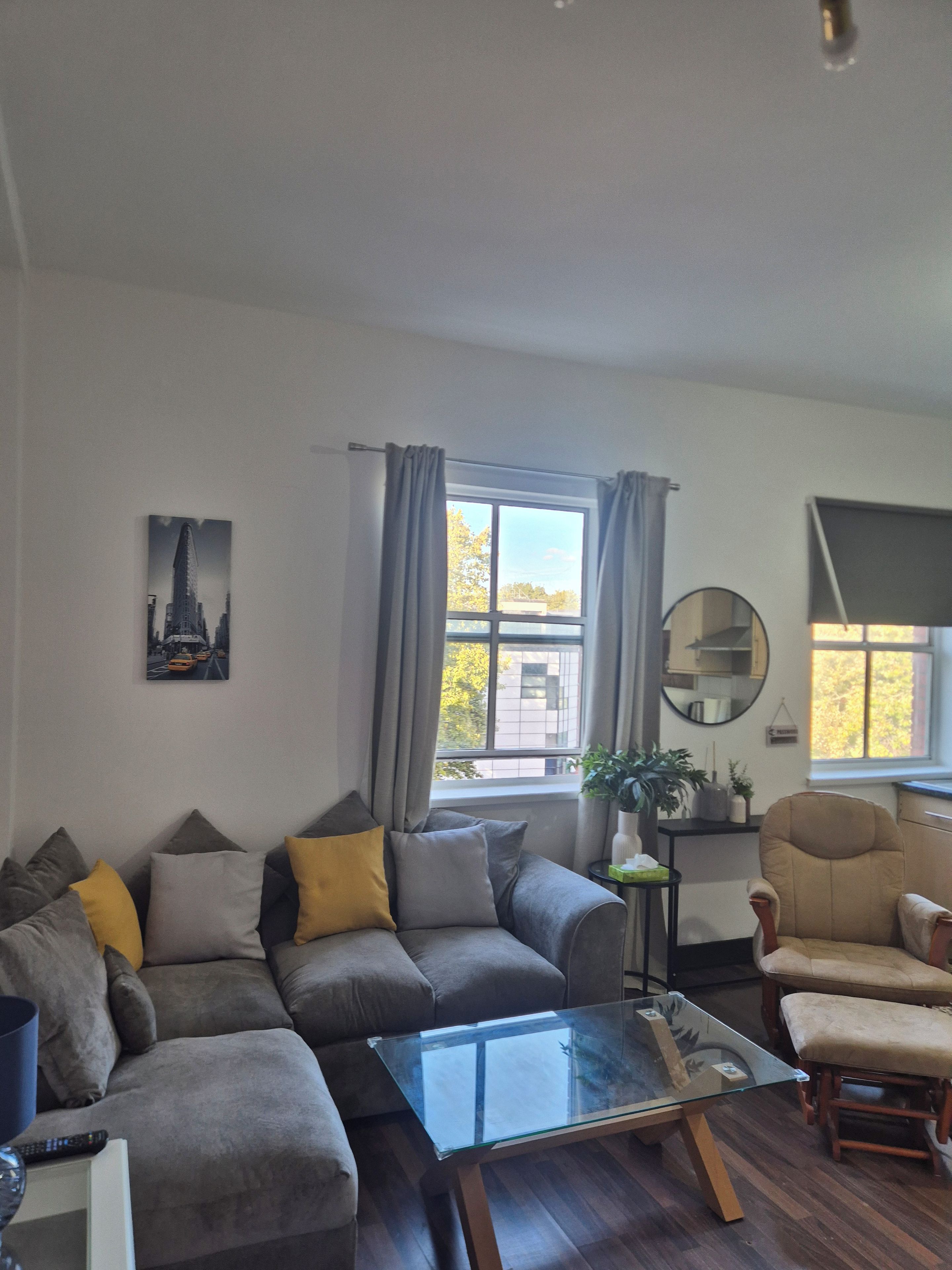 Een woonkamer met een hoekbank, een glazen salontafel, een ronde spiegel en uitzicht naar buiten door een raam.