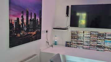 Una stanza con un dipinto di una veduta cittadina, una TV a schermo piatto, una pila di DVD e una luce al neon.
