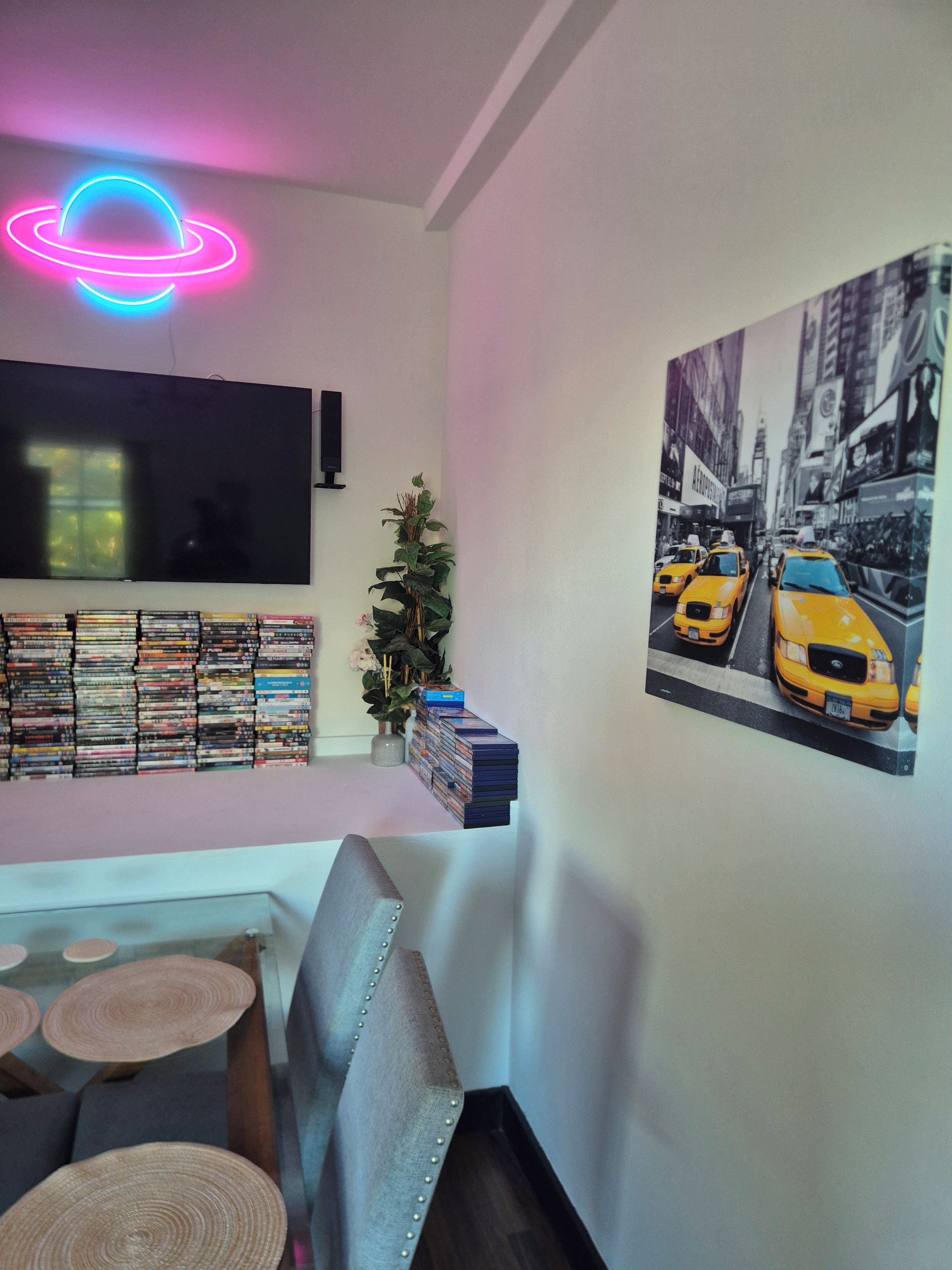 A living room with a flat-screen TV, a wall-mounted shelf with DVDs, a painting of taxis, and a neon light.