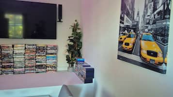 A living room with a flat-screen TV, a wall-mounted shelf with DVDs, a painting of taxis, and a neon light.