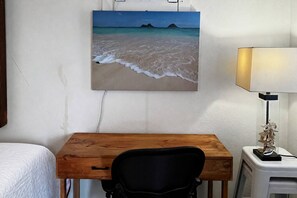 A room with a wooden desk, a chair, a lamp, and a painting of a beach scene on the wall. - Oceanfront Escape - Private Pier & Paddleboards (Kaneohe)