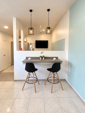 A modern kitchen with a central island, two black chairs, pendant lights, and a flat-screen TV mounted on the wall. - Paradise Studio Aruba 2 (Noord)