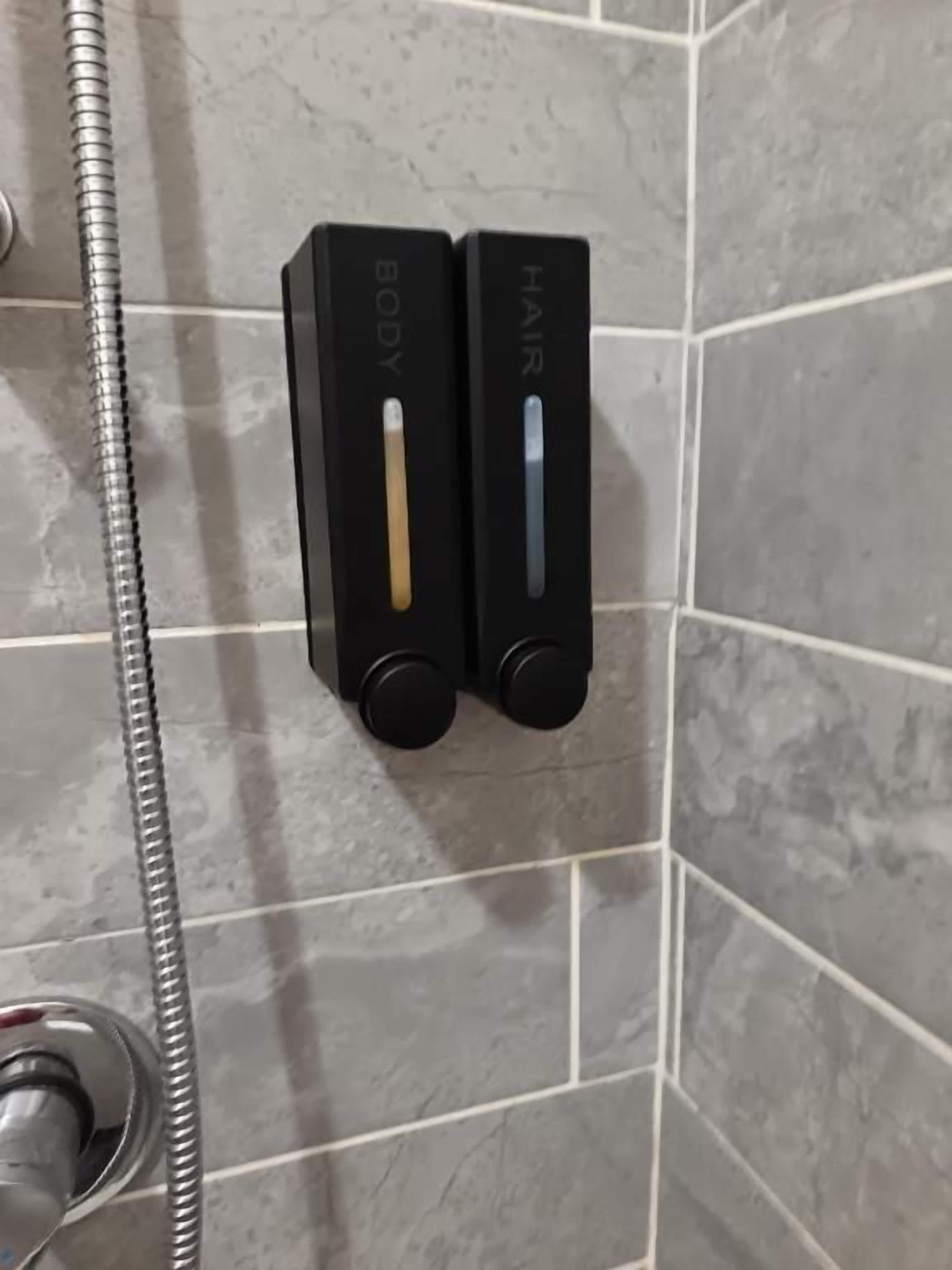 Area doccia con soffione a mano e due erogatori di sapone neri etichettati 'CORPO' e 'CAPELLI'.