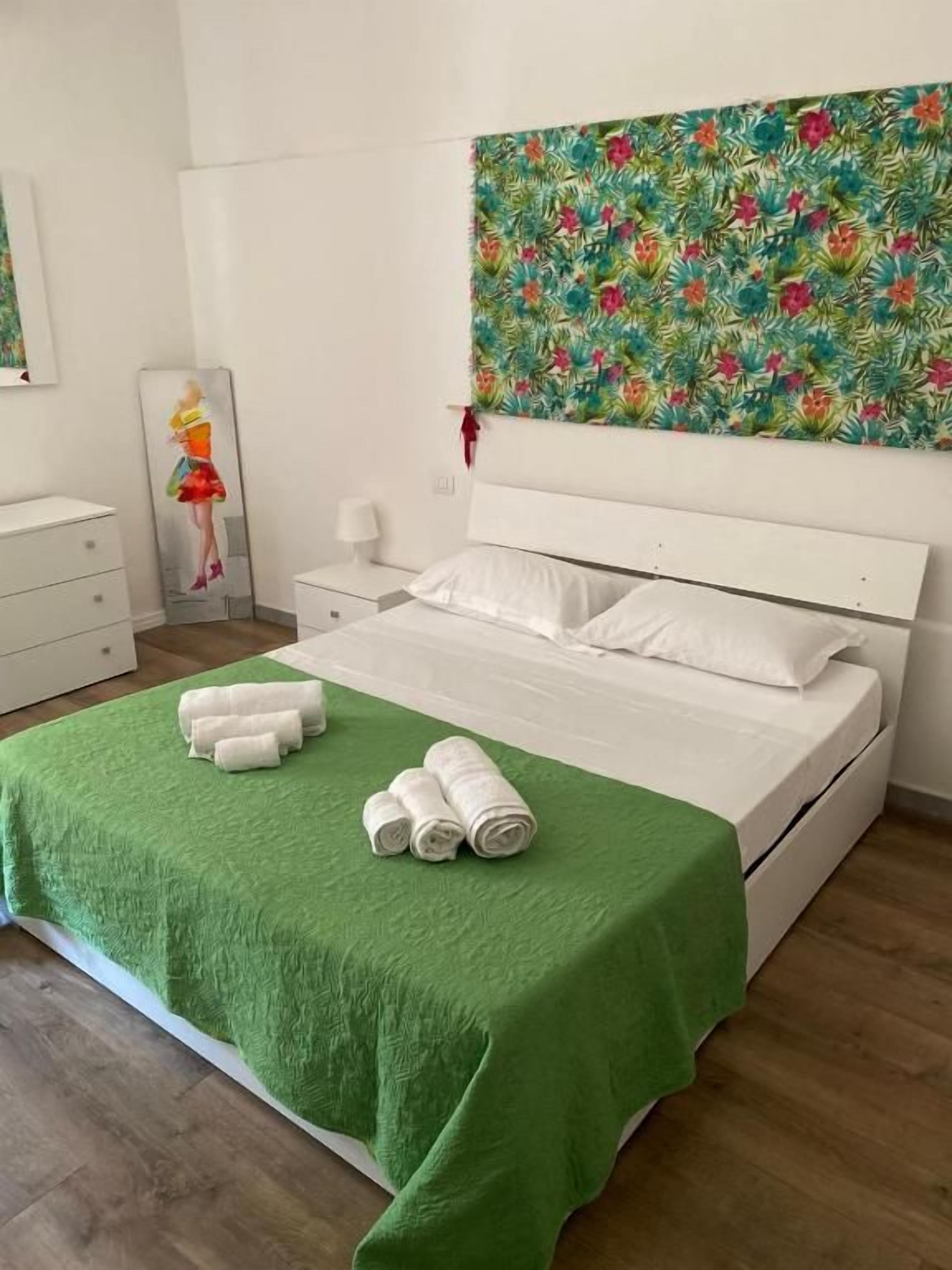 Un dormitorio con una cama, colcha verde, almohadas blancas, una mesita de noche, una lámpara y un cuadro en la pared.