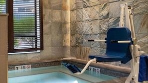Una pequeña piscina interior con pared de piedra, una ventana con persianas y un elevador de piscina.