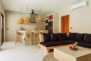 A modern kitchen with a dining area, a black sofa, and a coffee table with fruit. - Villa Pearl — Easy Island Living, Your Way (Ko Samui District)
