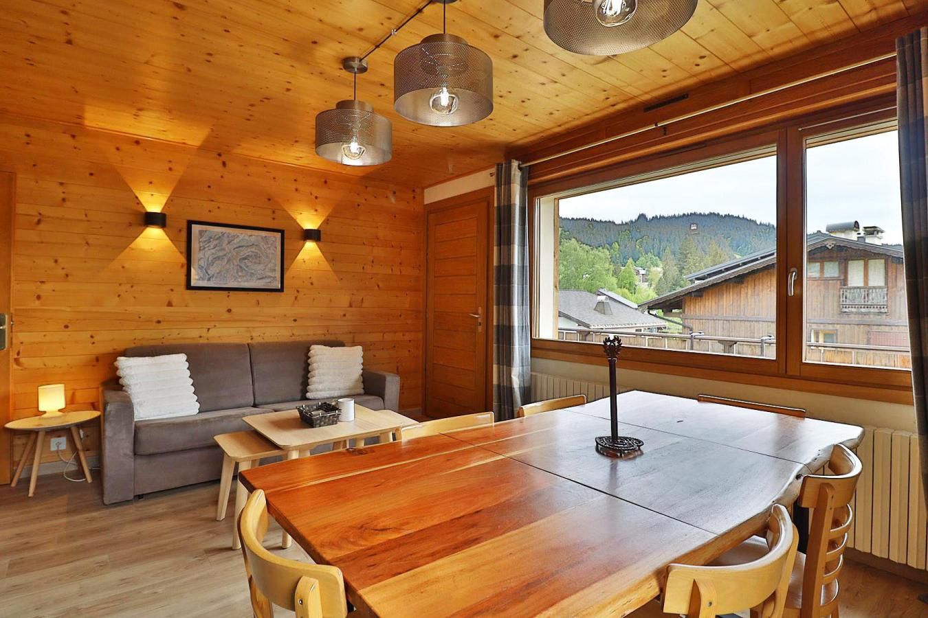 A wooden interior with a sofa, table, and chairs, a window with a view of buildings and hills, and pendant lighting.