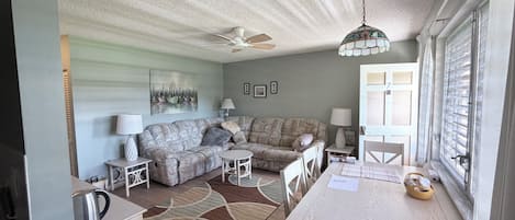 A living room with a sectional sofa, a dining table, a ceiling fan, and a window with blinds.