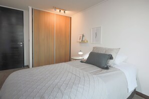 A bedroom with a bed, a bedside table with a lamp, a wardrobe, and a wall-mounted shelf with decorative items. - Modern 2BR Apartment in Las Condes (Las Condes)
