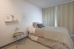 A bedroom with a bed, a desk with a lamp, a small shelf with items, and a window with curtains. - Modern 2BR Apartment in Las Condes (Las Condes)